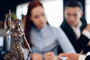Bronze statue of Themis holds scales of justice. In unfocused background, couple signs documents.